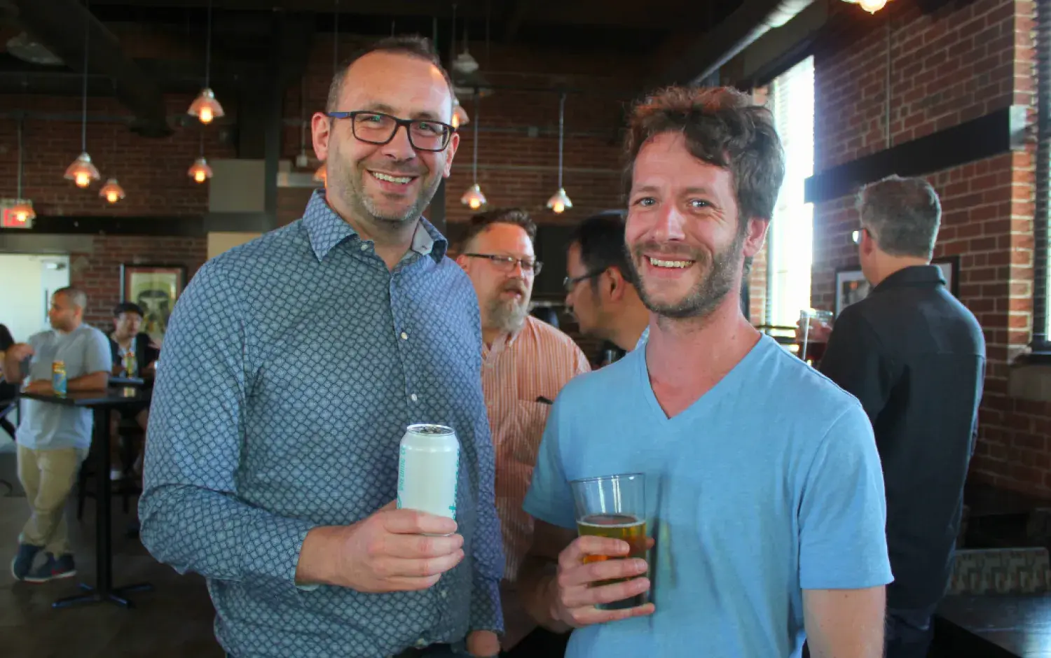 Two people holding drinks at a casual indoor gathering with brick walls and large windows.