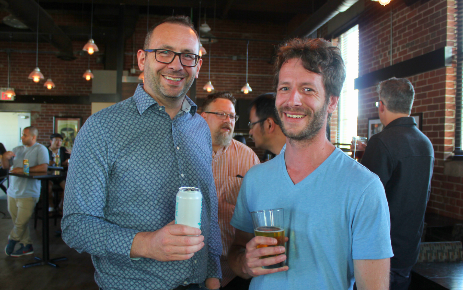 Two people holding drinks at a casual indoor gathering with brick walls and large windows.