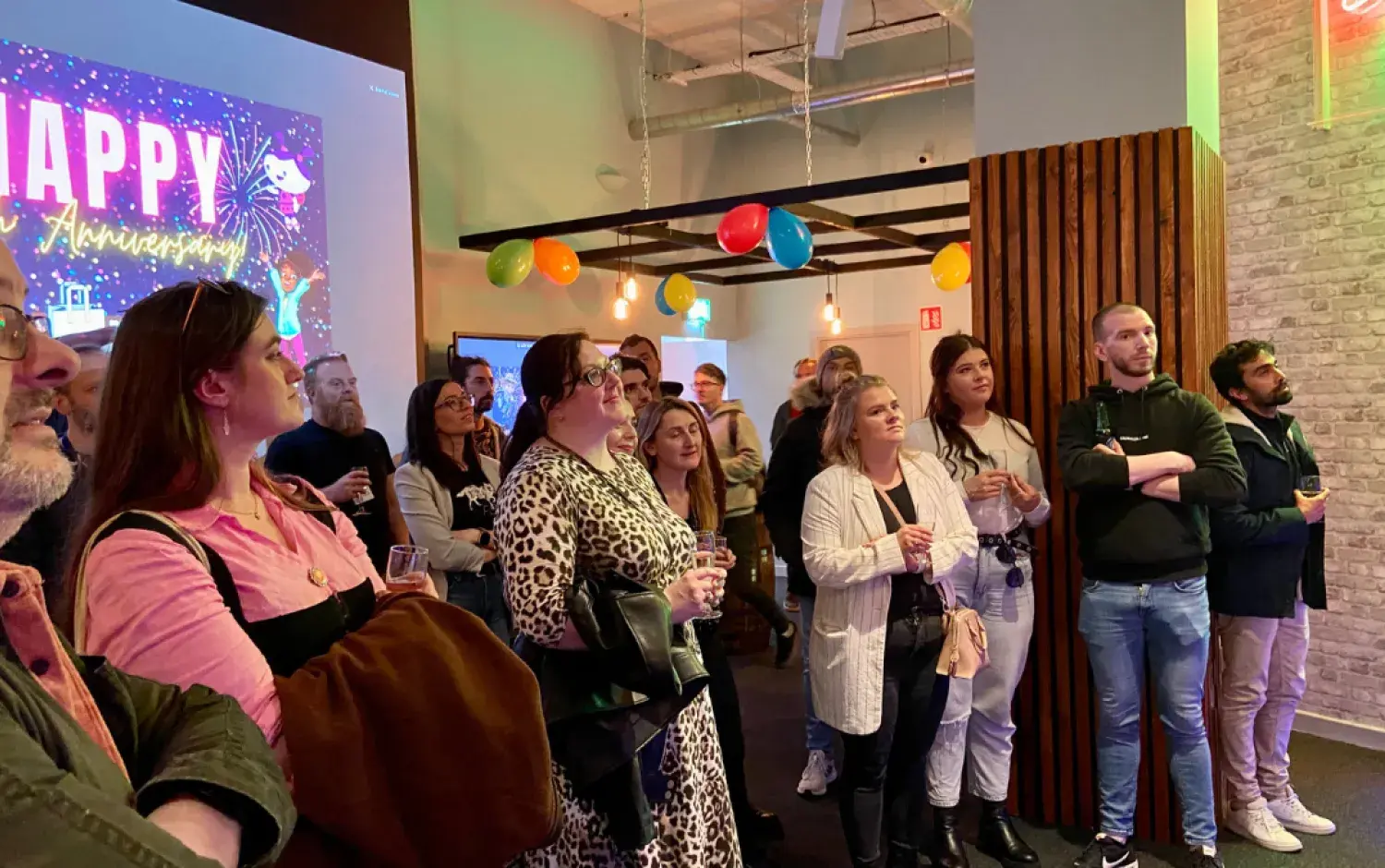 Group of people standing in a decorated room with balloons and a screen displaying “Happy Anniversary.”