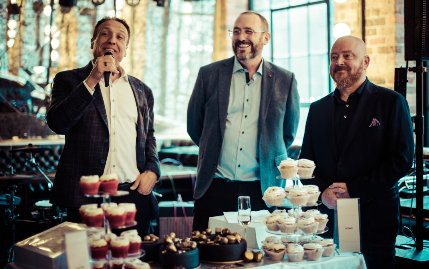 Three people standing behind a table filled with cupcakes and desserts at a celebration event.