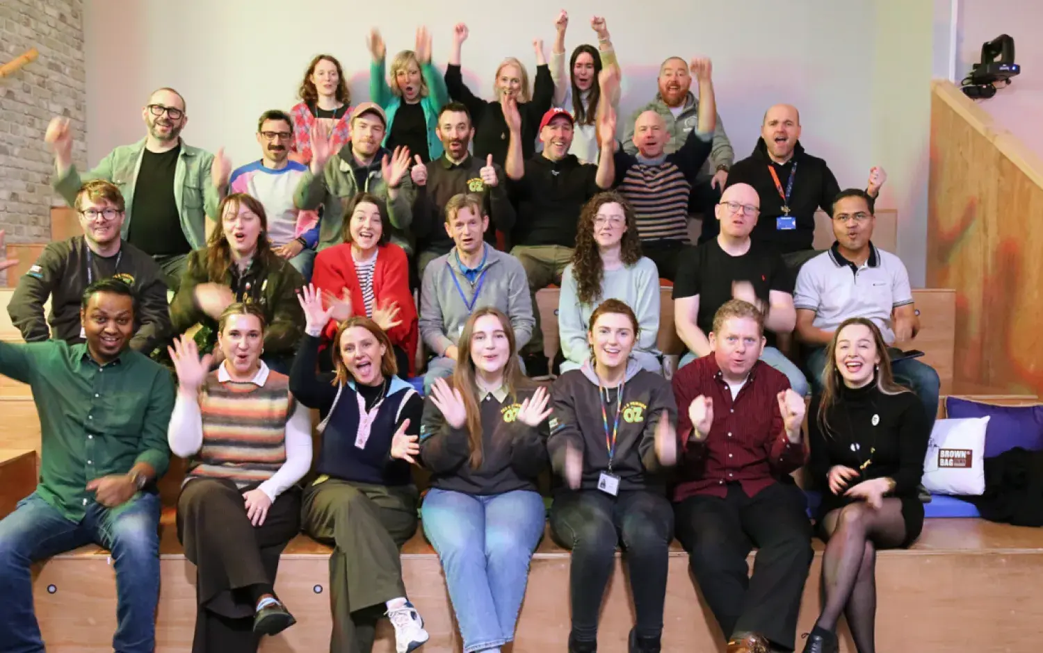 Large group photo of people seated on tiered steps indoors, raising hands in a celebratory pose.