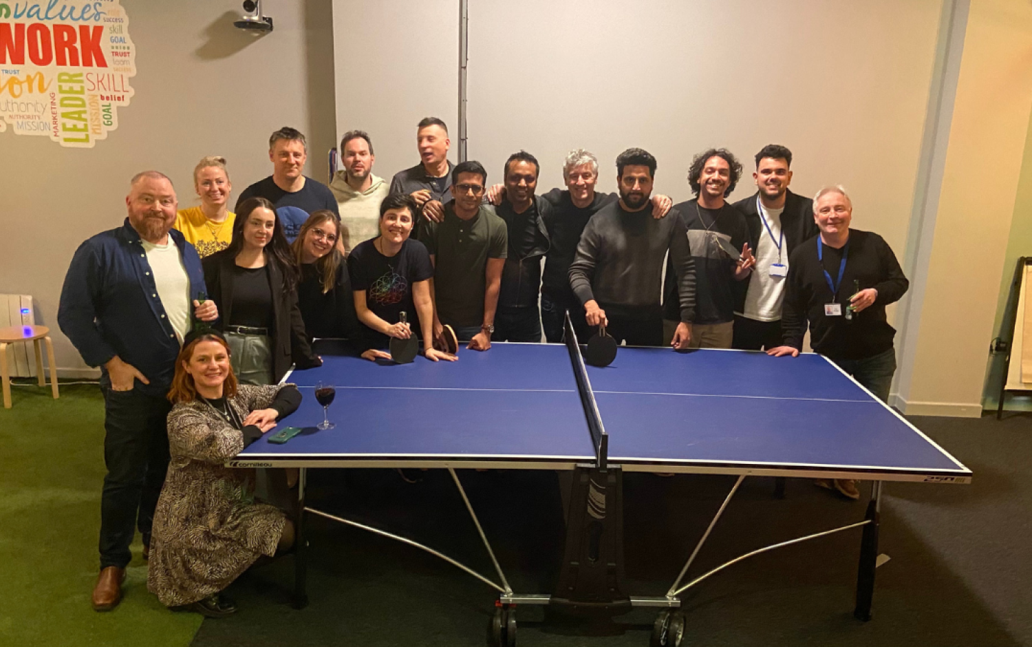 Group standing around a ping pong table indoors, some holding drinks during a casual office event.