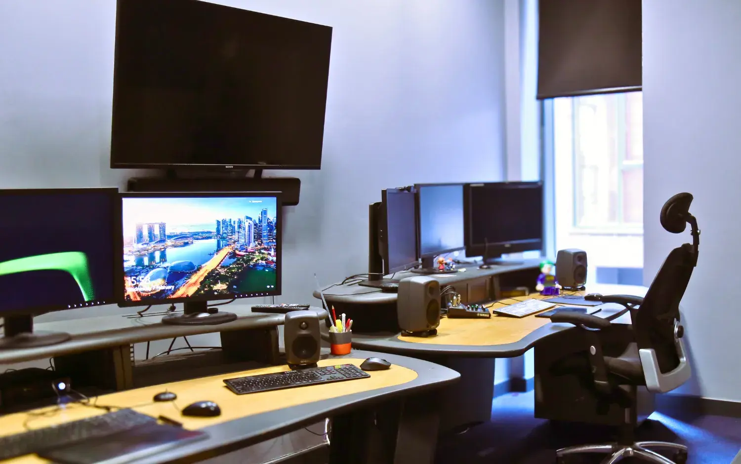 Modern office workspace with multiple computer monitors, large wall-mounted screen, ergonomic chair, speakers, and desks arranged in a bright room with a window.