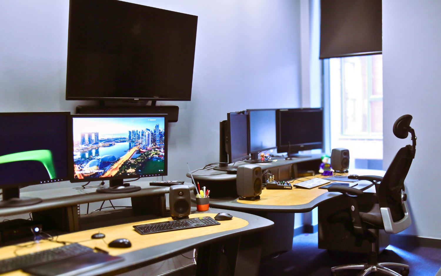 Modern office workspace with multiple computer monitors, large wall-mounted screen, ergonomic chair, speakers, and desks arranged in a bright room with a window.