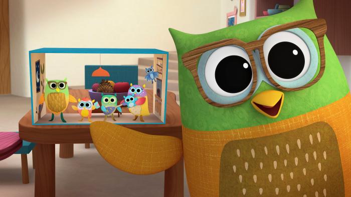 Green owl in front of a diorama with colorful paper cut outs of an owl family.